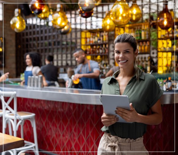A smiling woman holding a tablet in a festive bar setting.