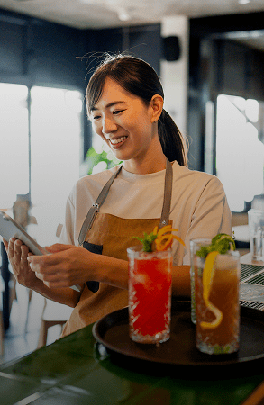 A woman smiling while holding a phone behind a bar with colorful drinks.